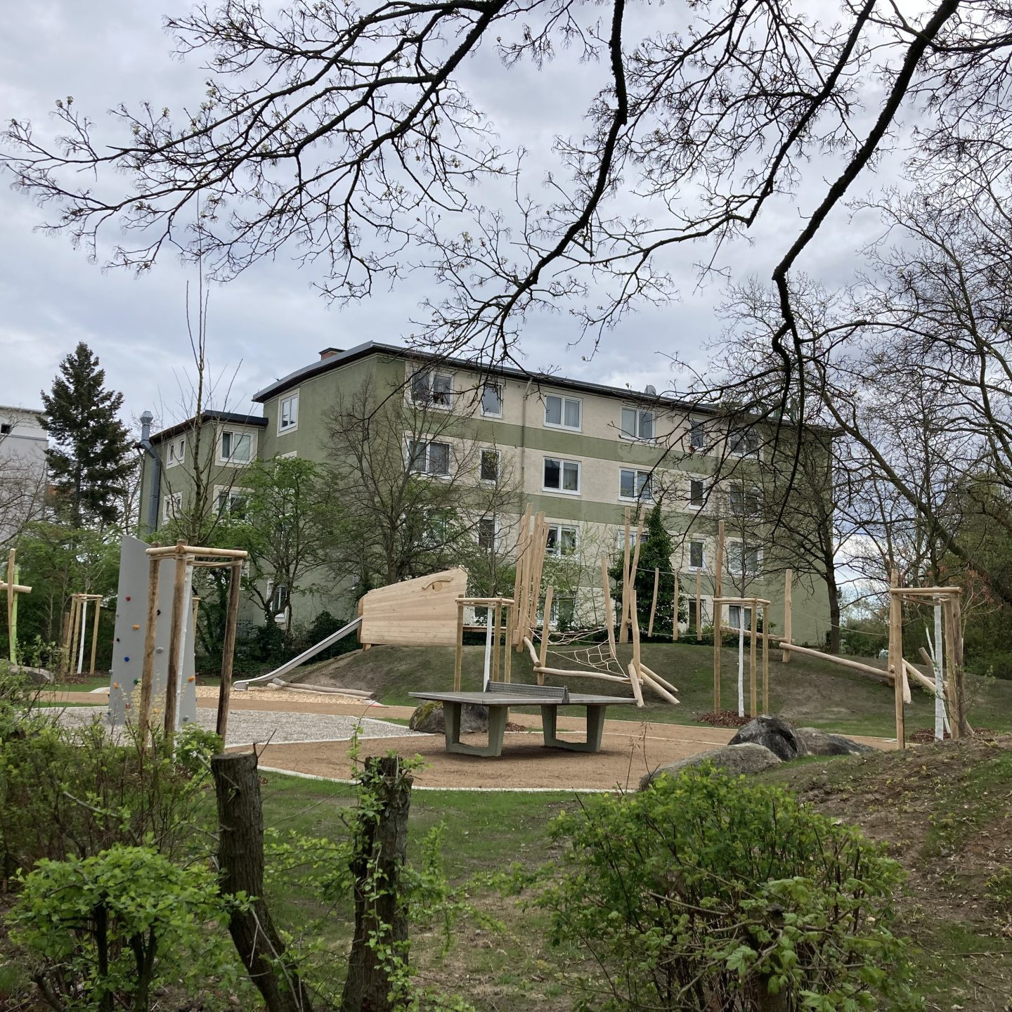 Spielplatz u.a. mit Schaukel, Kletterwand, TT-Platte und Mehrfamilienhäusern im Hintergrund