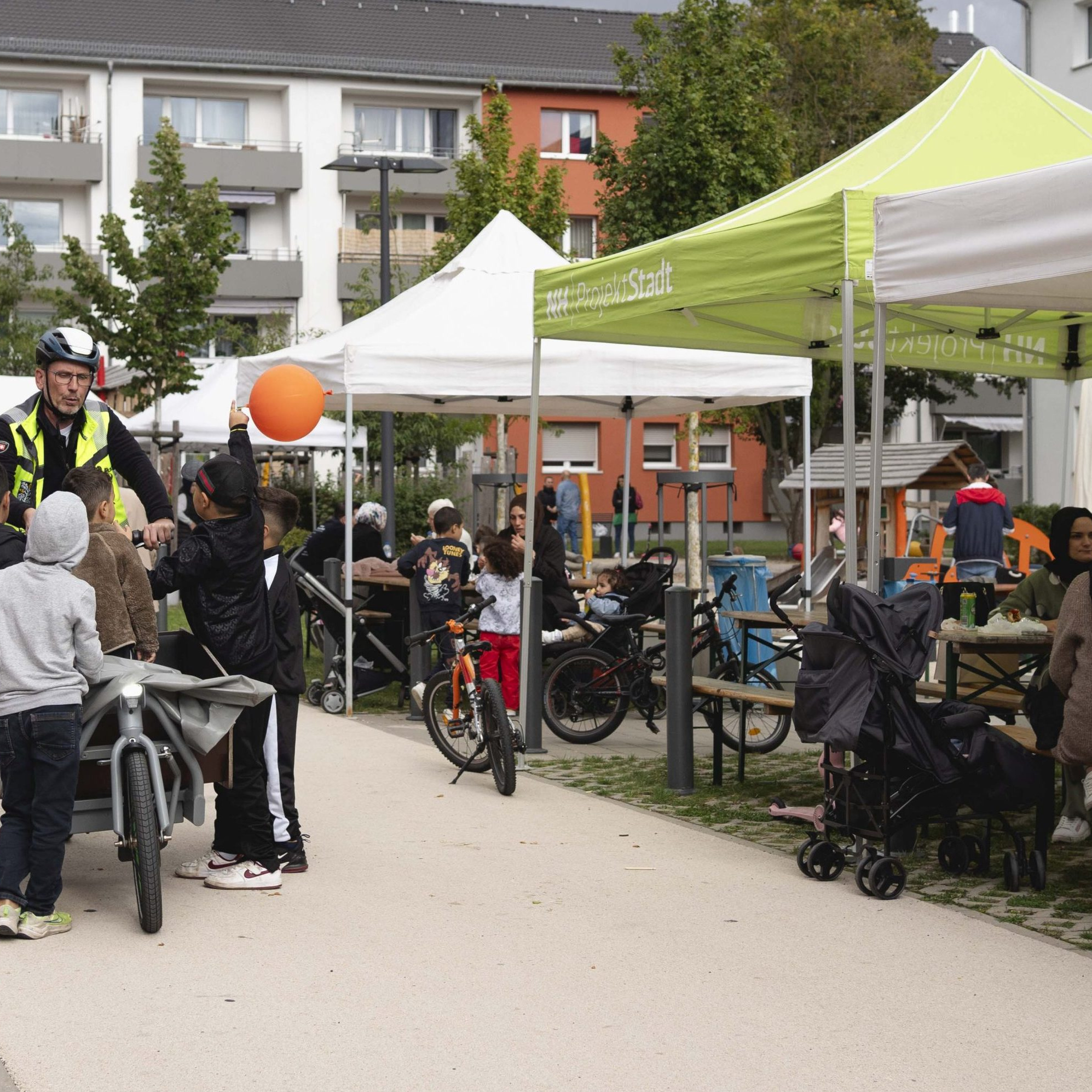 Ein Mann auf einem E-Lastenrad, umringt von Kindern, eines davon mit einem Luftballon, an der Seite Menschen, die an Tischen unter Pavillons sitzen.