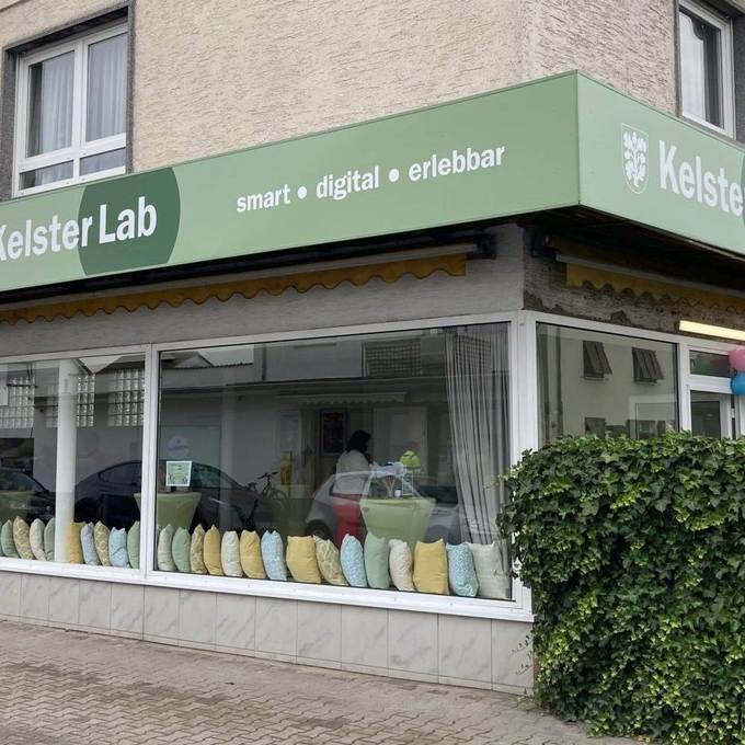 Das KelsterLab von außen, mit Sitzkissen in den Schaufenstern, Hecke am rechten Bildrand