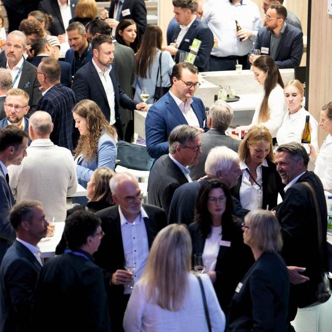 Viele Menschen, alle schick gekleidet, stehen an einem Messestand und unterhalten sich miteinander