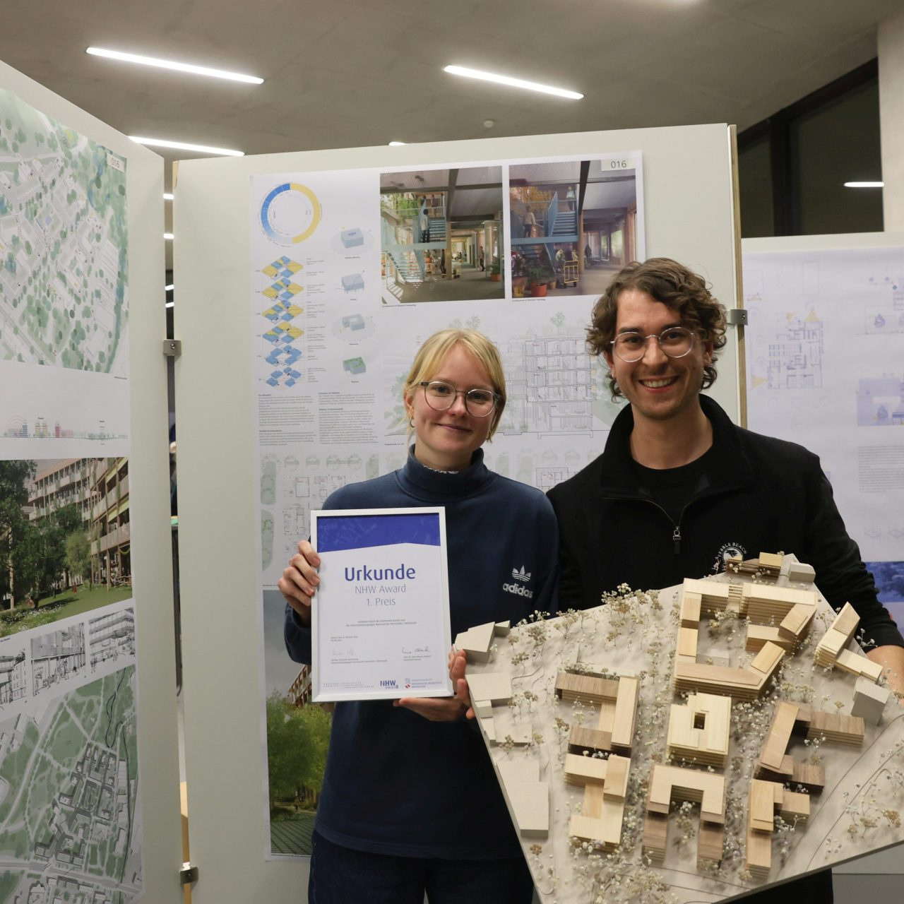 Elina Klett und Tom Stehmann belegten mit ihrem Projekt Klimahof einen der beiden ersten Plätze. Urkunde und Modell halten Sie in den Händen, im Hintergrund ihre Arbeit auf einer Plakatwand