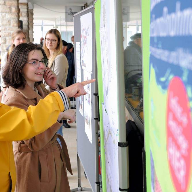 Zwei Frauen stehen vor einem Plakat und zeigen mit den Fingern darauf